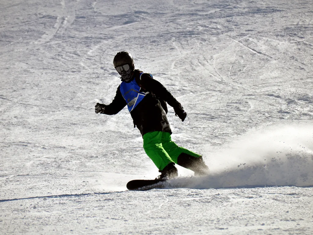 Snowboarder in grüner Hose bremst mit Schneefontäne.